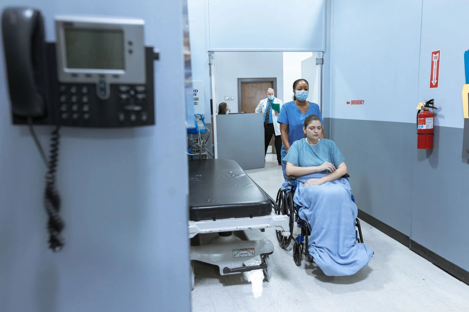 Couloir d'un service d'urgences hospitalier, patiente en fauteuil roulant accompagnée par une infirmière